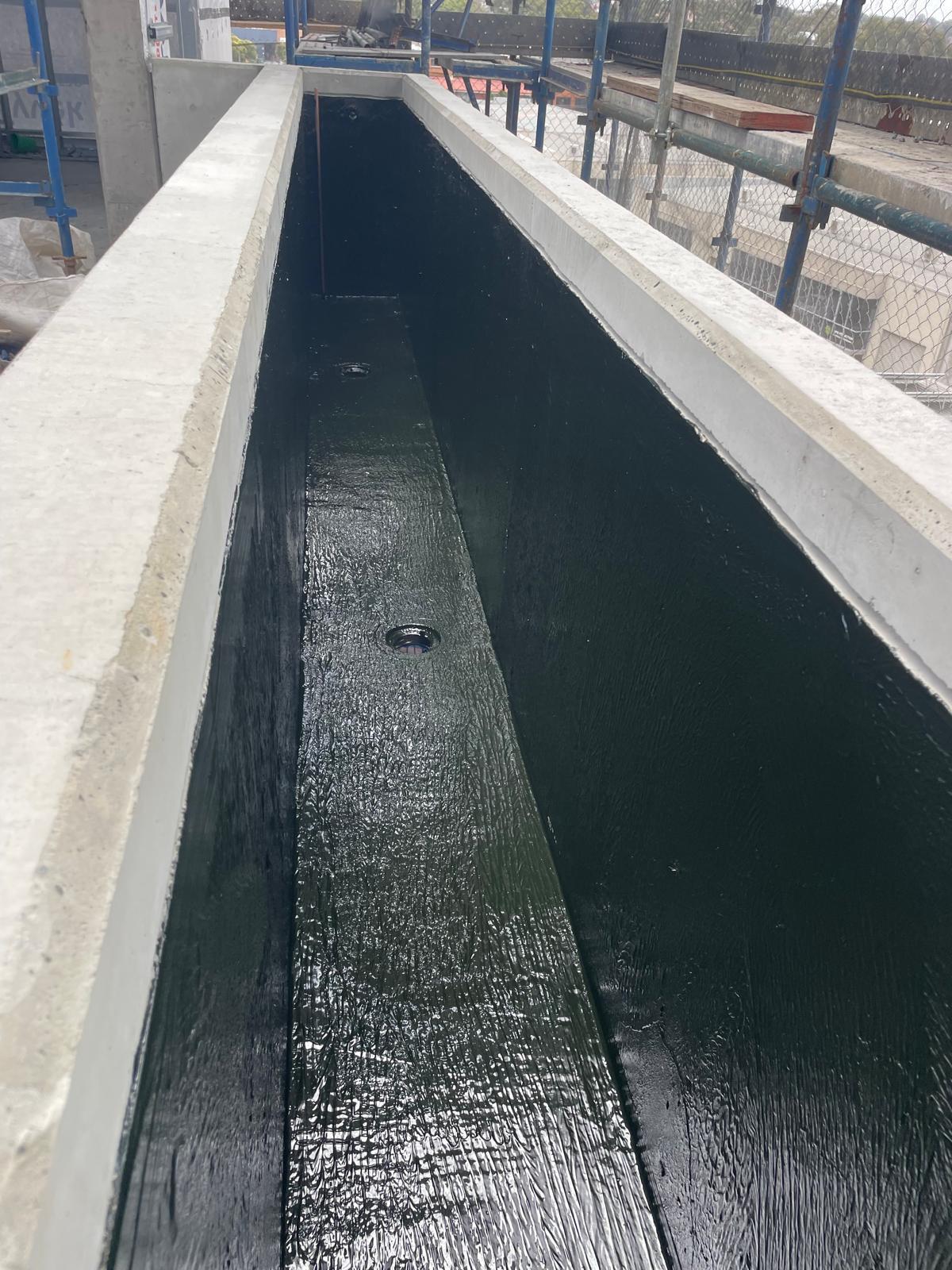 Long narrow concrete planter box lined in shiny black membrane with two drainage outlets