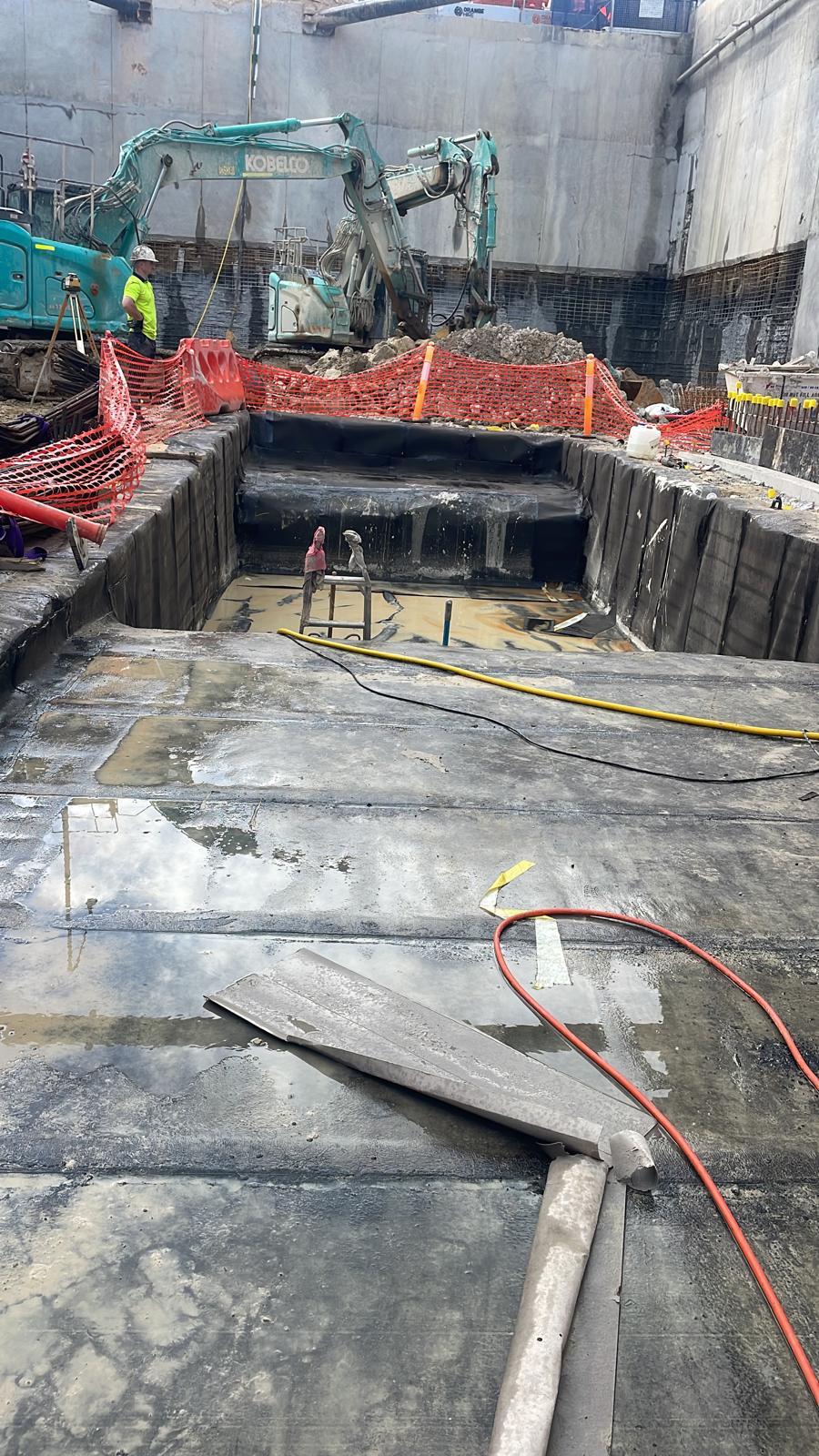 Lift pit excavation with walls and floor lined in black tanking membrane beside an excavator