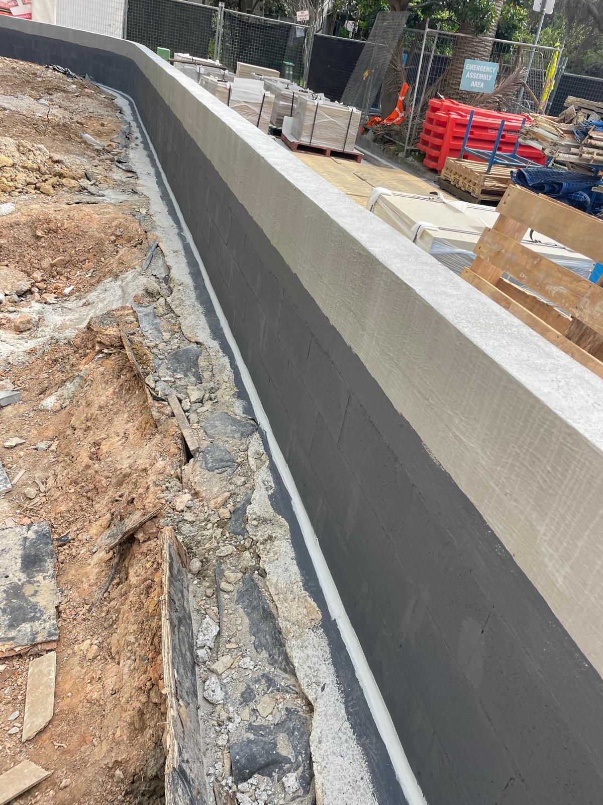 Curving block retaining wall partially coated in dark grey membrane beside a dirt trench