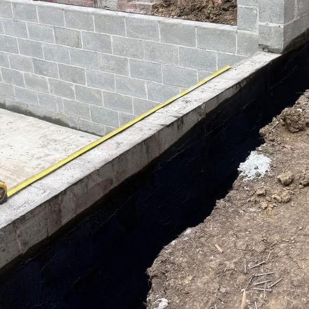 Concrete retaining wall footing tanked in black membrane before backfill, with a tape measure across the top
