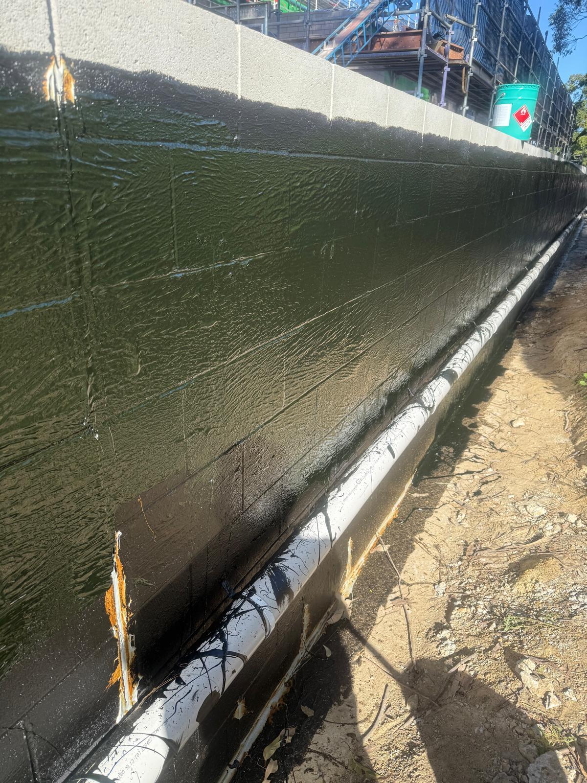 Dark green liquid membrane covering a long block wall with a white ag pipe along the footer