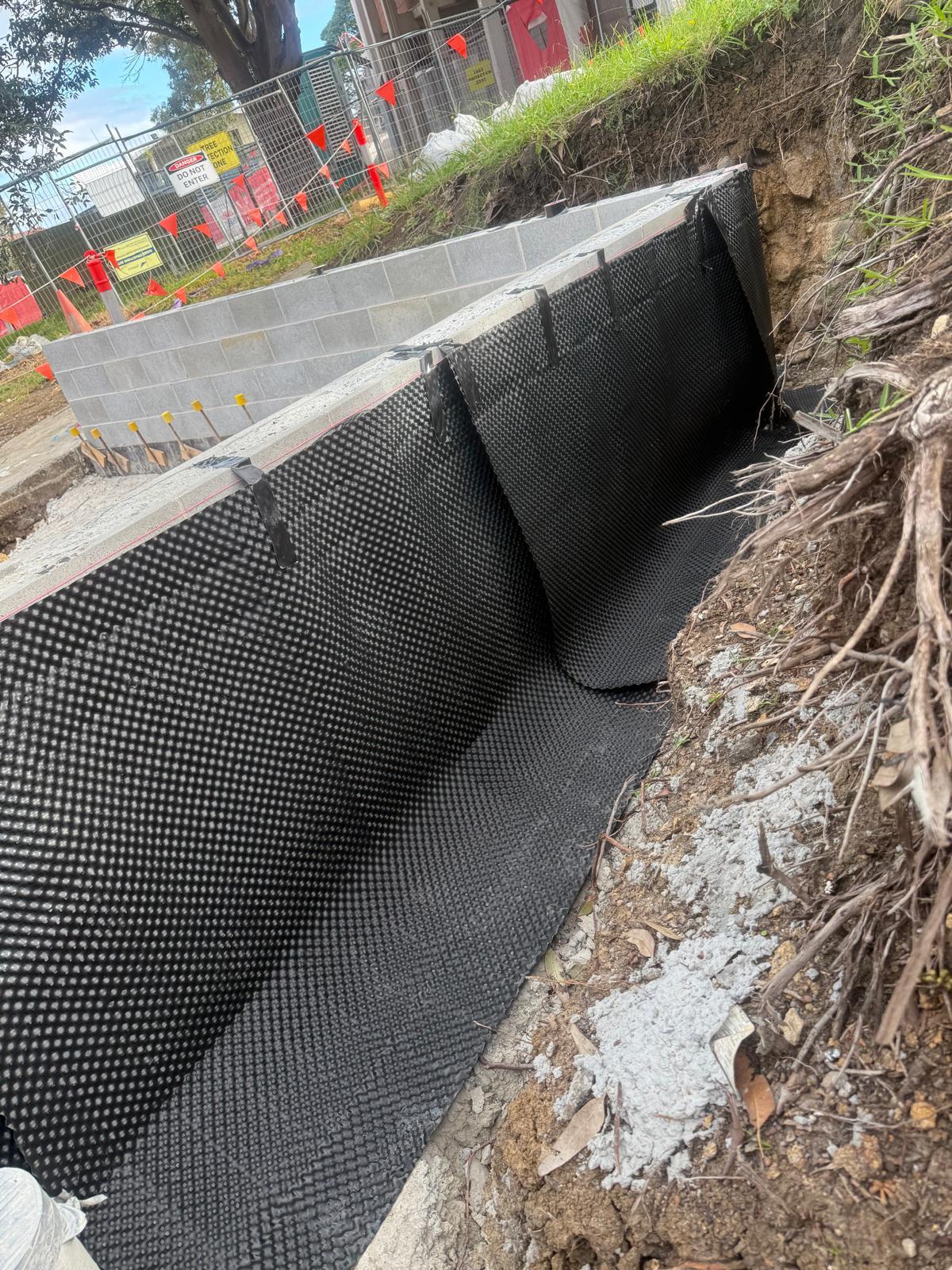 Black dimpled sheet membrane installed against the back of a block wall inside an excavated trench