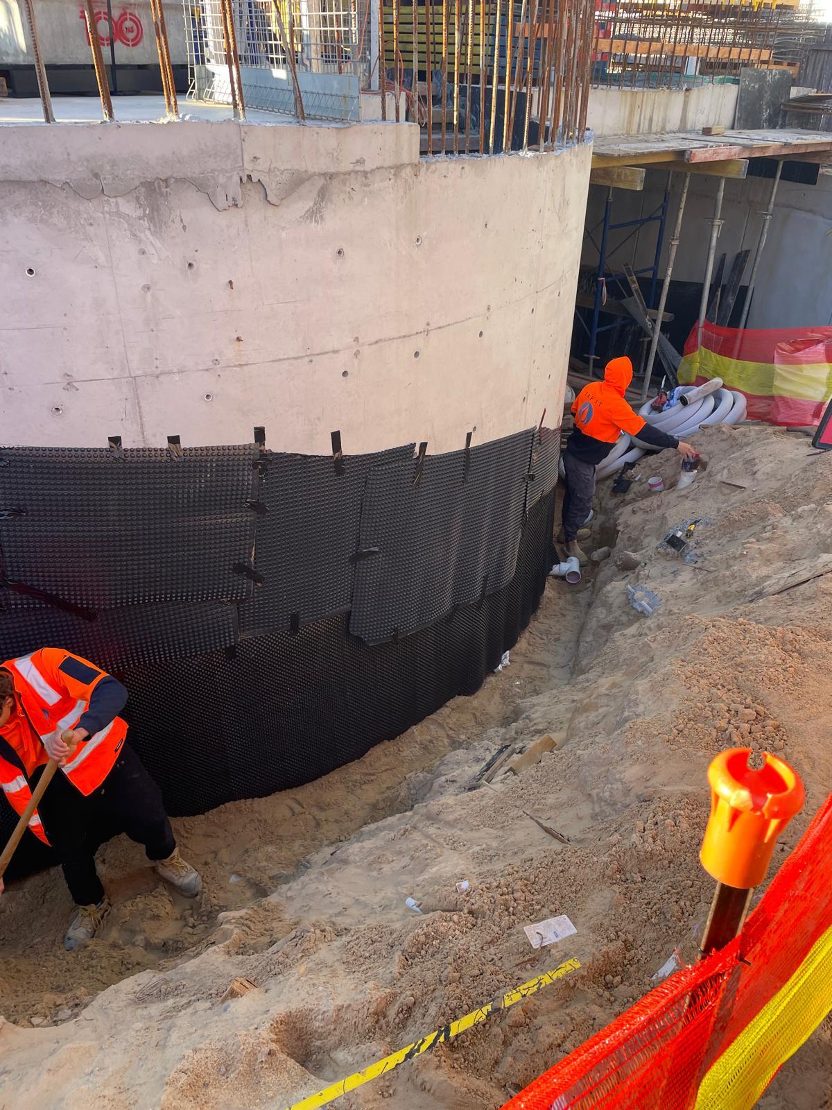 Workers in orange gear installing black dimpled drainage board over a waterproofed curved wall