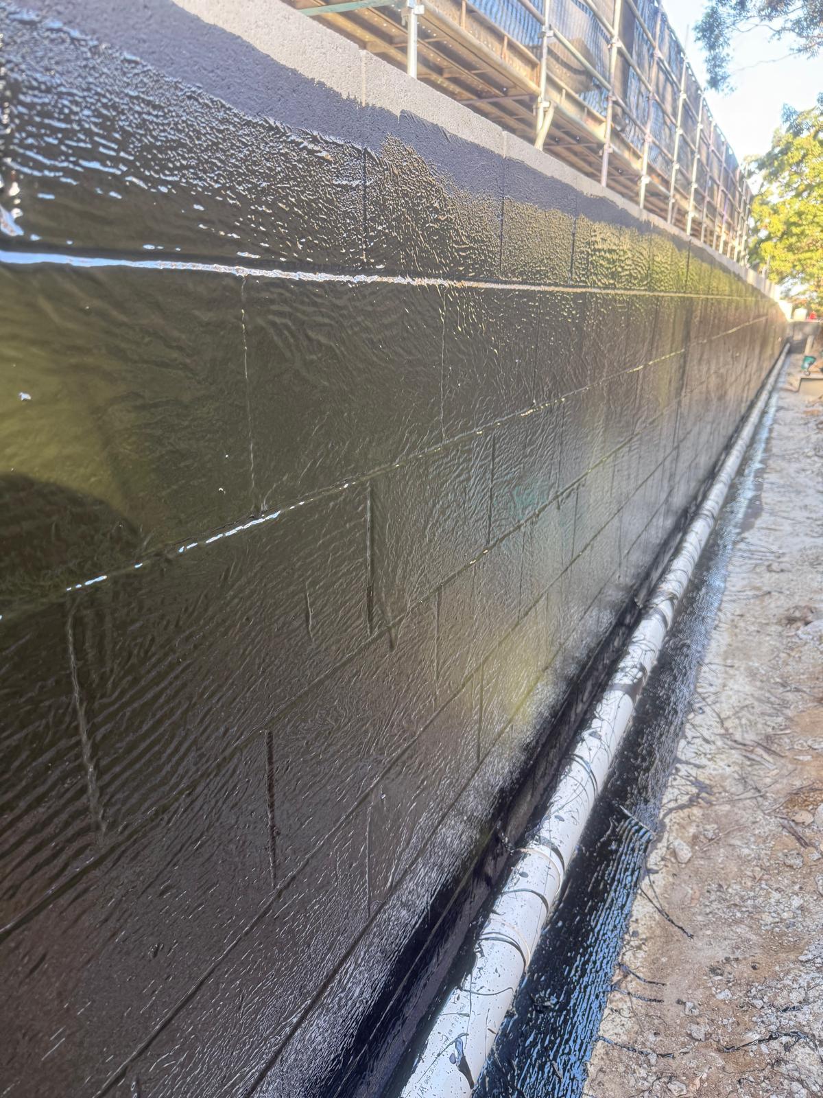 Glossy dark brown membrane applied to a block retaining wall with a white PVC drain pipe at the base