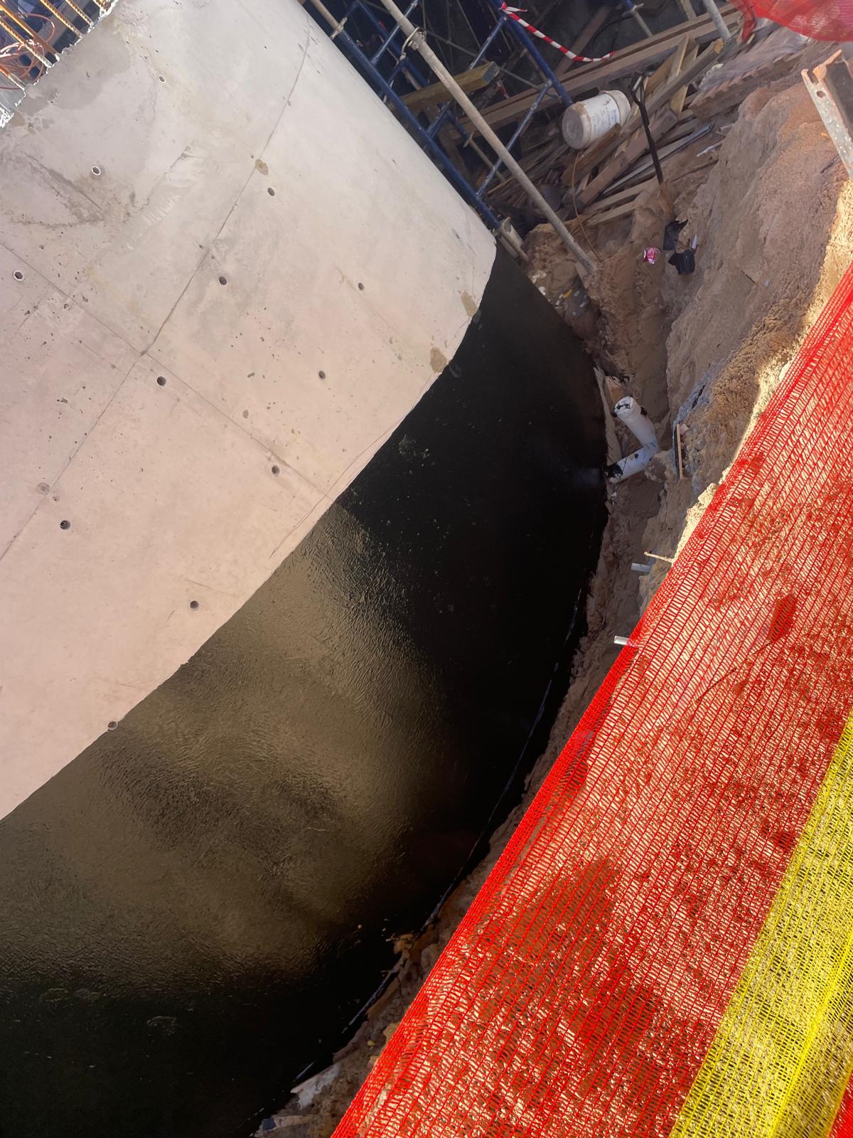 Curved concrete retaining wall with black liquid membrane on its lower section beside an open trench