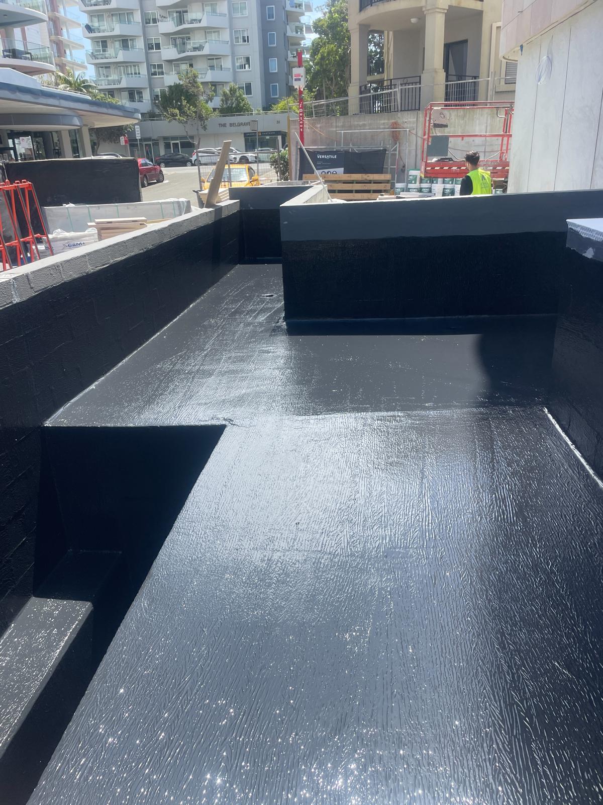 Multi-angled concrete planter box coated in shiny black waterproofing membrane on an urban site