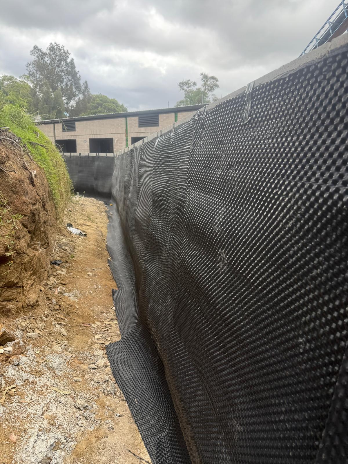 Black dimpled drainage sheet installed the full height of a tall retaining wall in an excavated trench