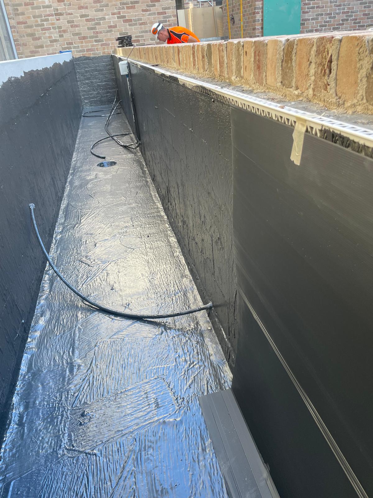 Long narrow planter box lined with dark grey membrane with a central drain and black conduits